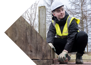 A student in a safety vest kneels down, placing bricks on a wooden structure amidst a backdrop of trees and a cloudy sky.