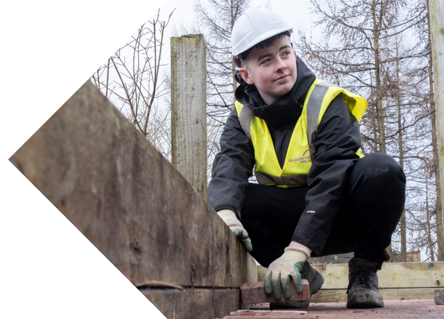 A student in a safety vest kneels down, placing bricks on a wooden structure amidst a backdrop of trees and a cloudy sky.