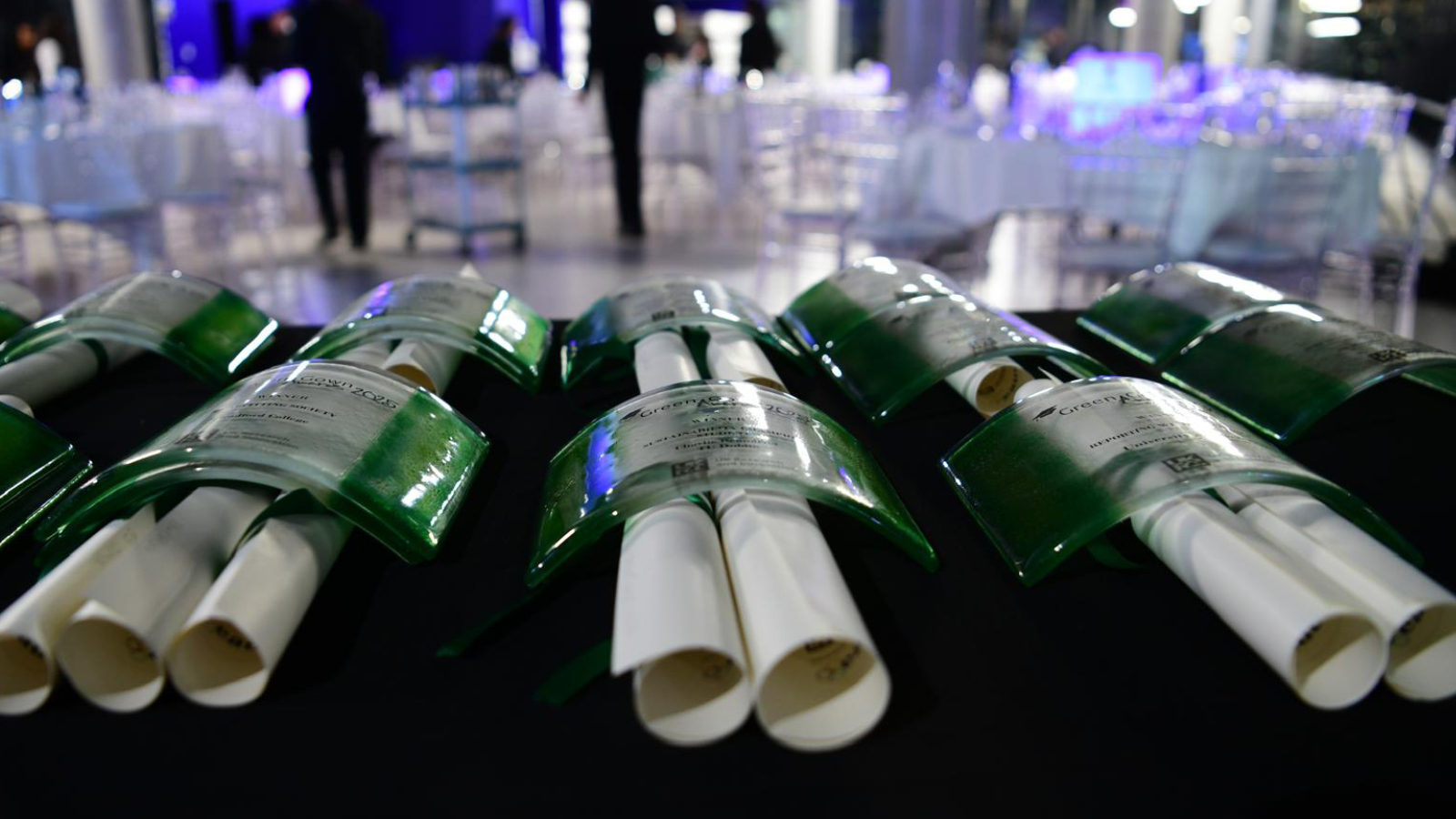 A display of awards featuring glossy green plaques over rolled certificates, set against a softly lit reception backdrop.