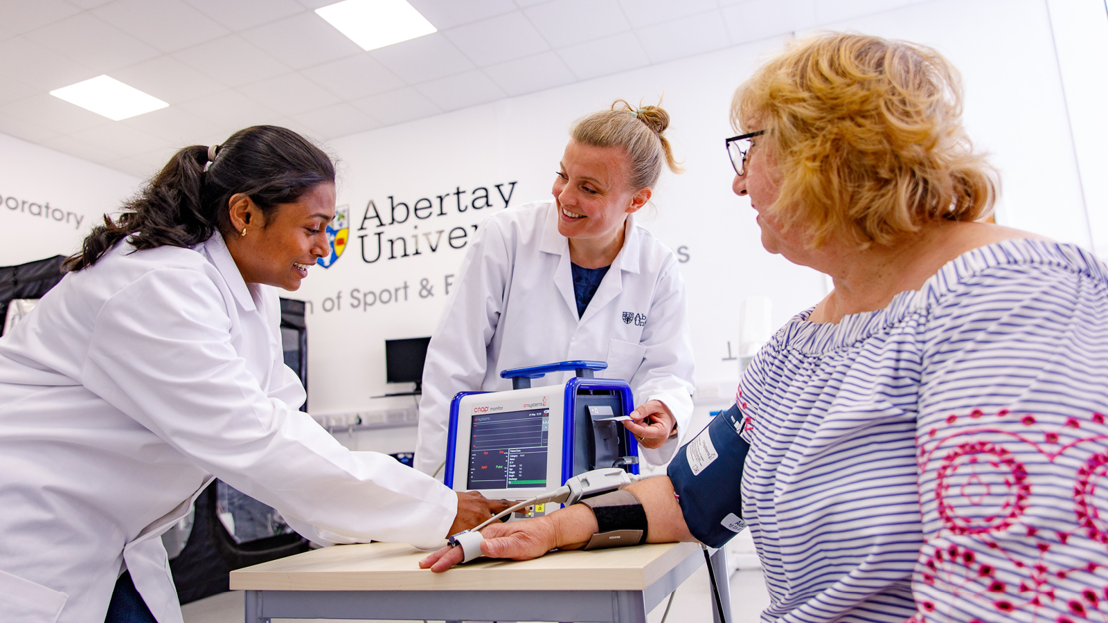 Two researchers in lab coats demonstrate a medical test using a monitoring device on a participant’s arm.