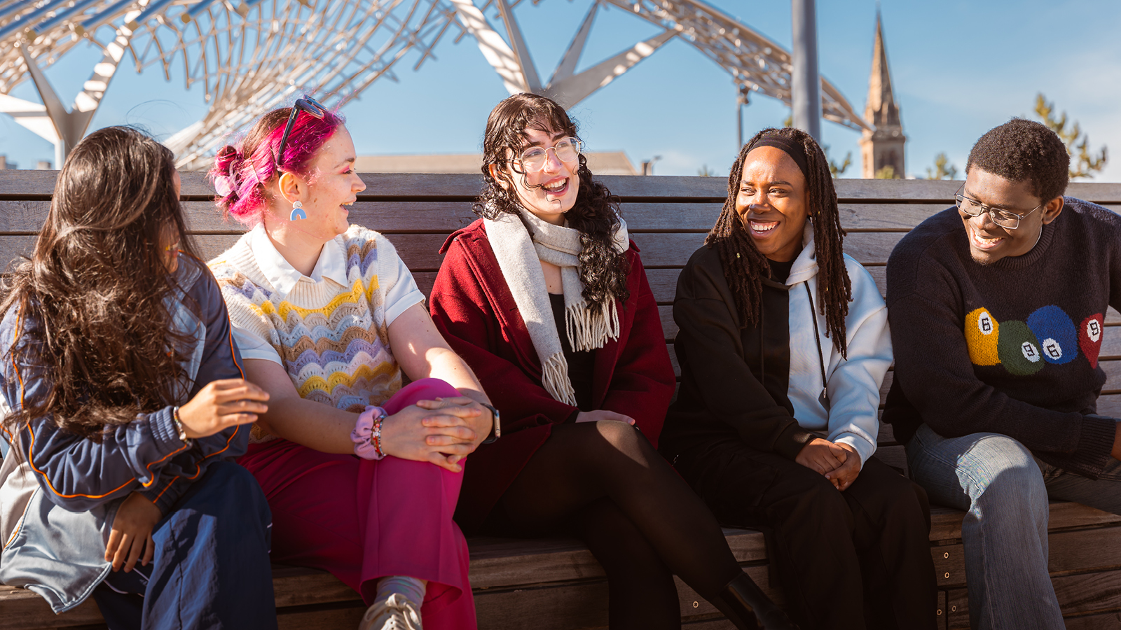 Group of five people sit on a bench outdoors, chatting together with a modern structure behind them.