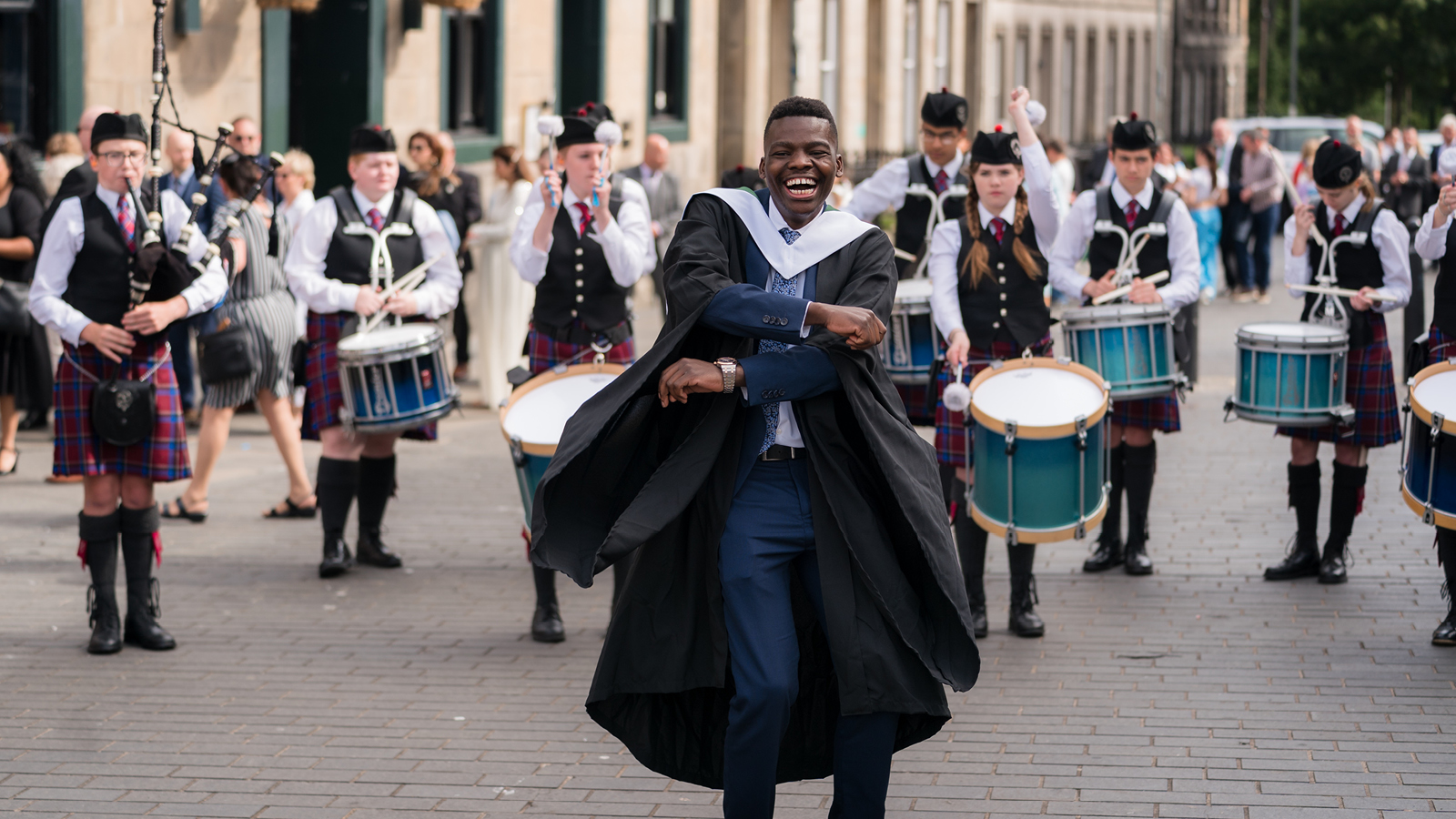 Graduate in academic gown walking in front of a pipe band during a graduation procession on a city street.